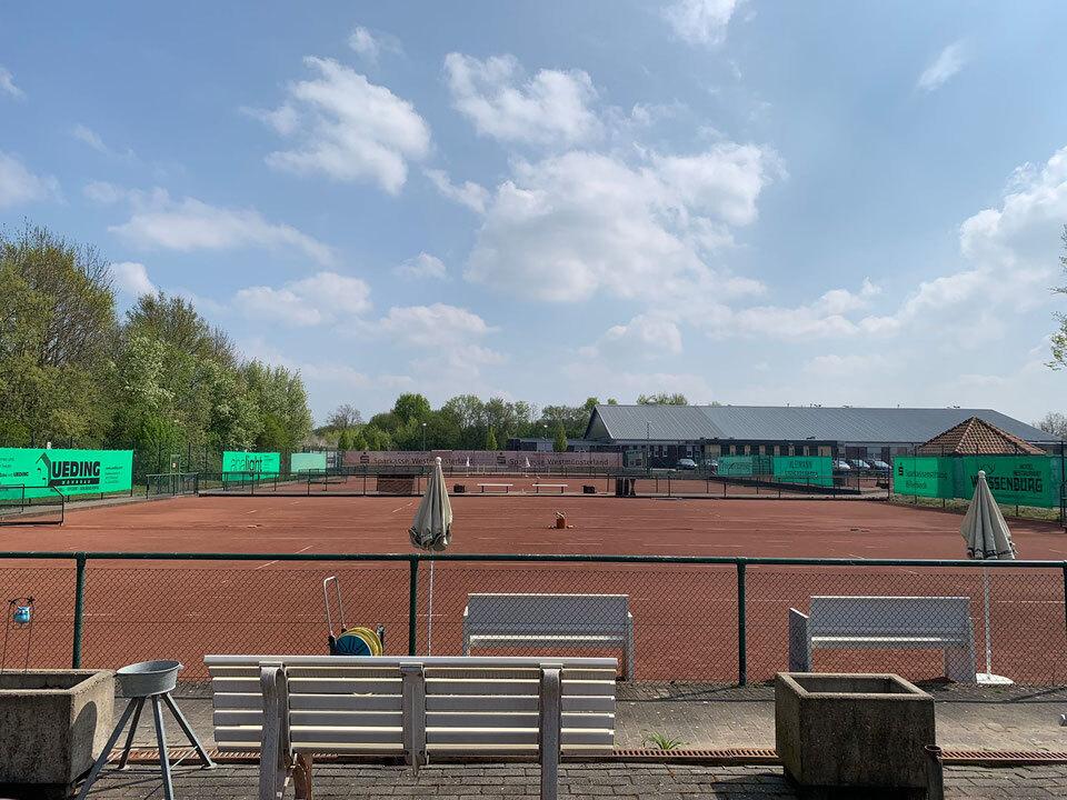 Panoramaaufnahme eines Tennisplatzes mit rotem Sand, Bänken und schattenspendenden Schirmen unter einem blauen Himmel.