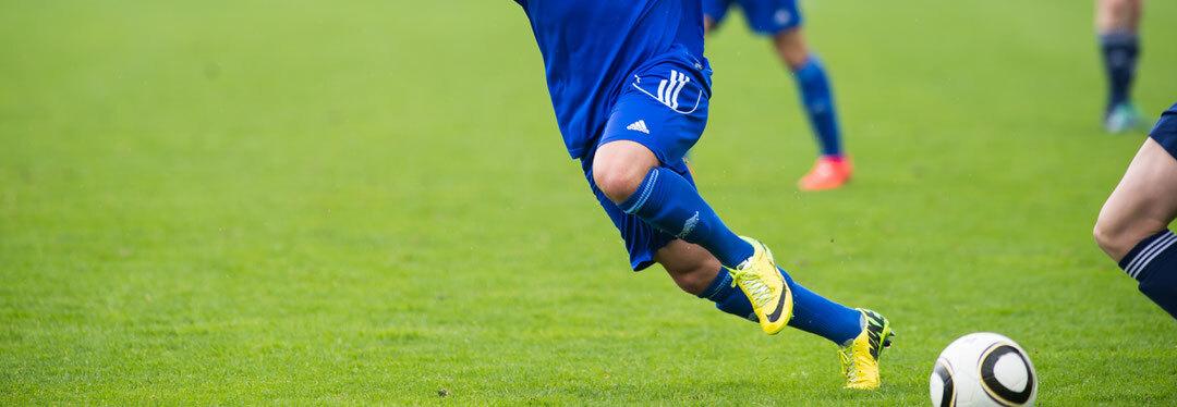 Spieler in blauer Fu&szlig;ballausr&uuml;stung sprintet &uuml;ber den gr&uuml;nen Rasen, w&auml;hrend ein Fu&szlig;ball neben ihm rollt.