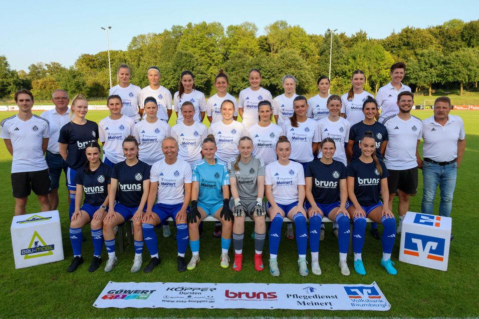 Teambild der Frauenfu&szlig;ballmannschaft mit Trainern, im Hintergrund ein gr&uuml;ner Rasenplatz und Werbebanner.