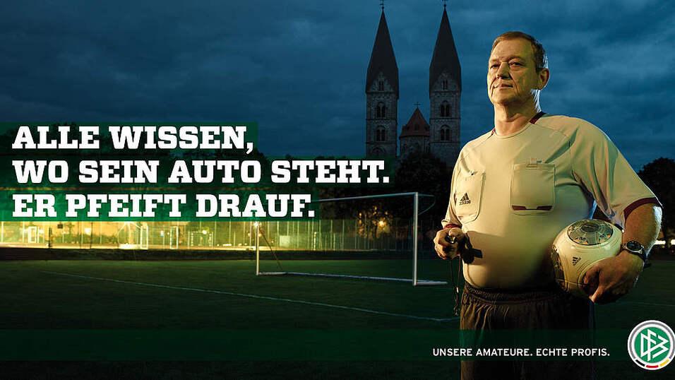 Schiedsrichter mit Pfeife und Ball auf einem Fu&szlig;ballplatz, im Hintergrund zwei Kircht&uuml;rme und ein bew&ouml;lkter Himmel.