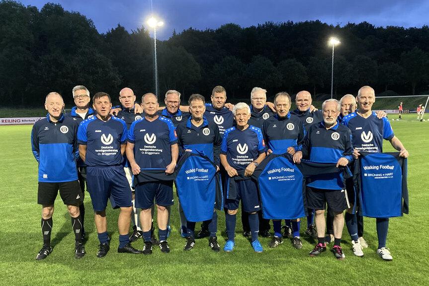Gruppe von 15 Männern in blauen Sportanzügen, die auf einem Fußballplatz stehen und T-Shirts mit "Walking Football" halten.