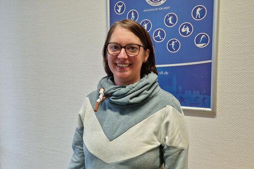 Frau mit Brille in grauem Sweatshirt steht vor einem Sportplakat an einer wei&szlig;en Wand.