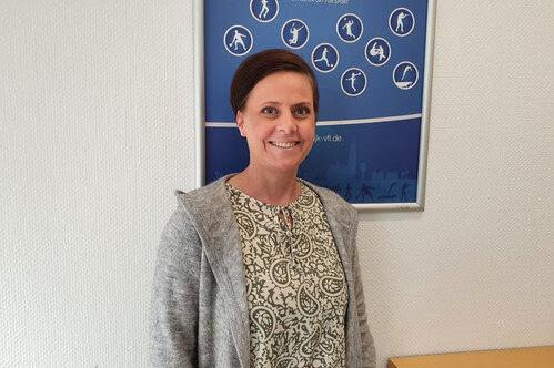 Frau mit kurz geschnittenen Haaren steht vor einem Sport-Poster in einem hellen Raum.