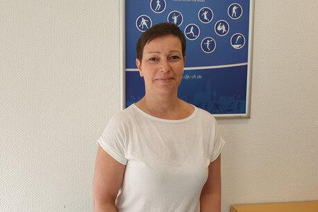 Frau mit kurzen Haaren in wei&szlig;em T-Shirt steht vor einem blauen Plakat mit Symbolen sportlicher Aktivit&auml;ten.
