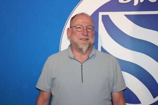 Mann mit Brille und kurzer Behaarung steht vor blauem Hintergrund mit einem Vereinslogo.