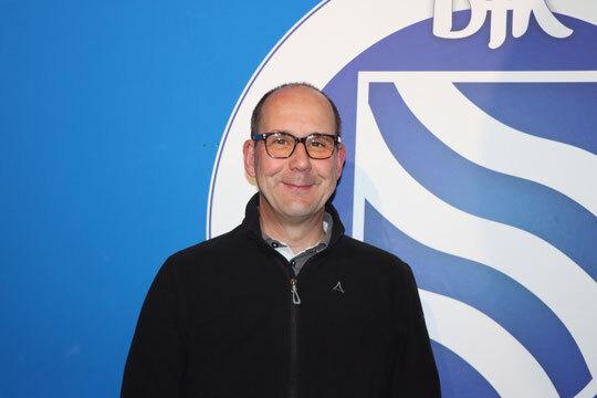 Mann mit Brille steht vor einer blauen Wand mit einem wei&szlig;en Logo in Wellenoptik.