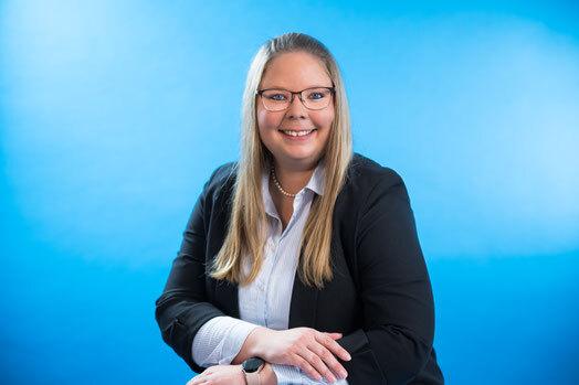 Portr&auml;t einer l&auml;chelnden Frau mit Brille, die in einem schwarzen Blazer vor einem blauen Hintergrund sitzt.