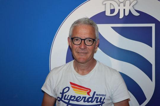 Mann mit Brille und grauen Haaren tr&auml;gt ein T-Shirt mit "Superdry", vor einem blauen Hintergrund mit Logo.