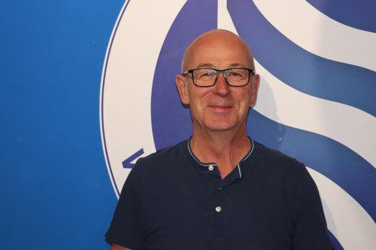 Portr&auml;t eines l&auml;chelnden Mannes mit Brille vor einem blauen Hintergrund mit geschwungenen wei&szlig;en Linien.