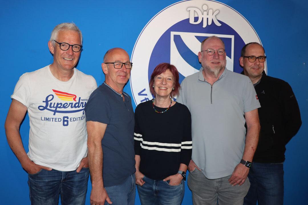 F&uuml;nf Personen posieren vor einer blauen Wand mit dem Logo eines Vereins. Alle l&auml;cheln und stehen nahe beieinander.