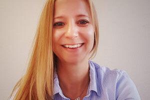 Portr&auml;t einer l&auml;chelnden Frau mit langen, blonden Haaren, die ein blaues Hemd tr&auml;gt, vor neutralem Hintergrund.