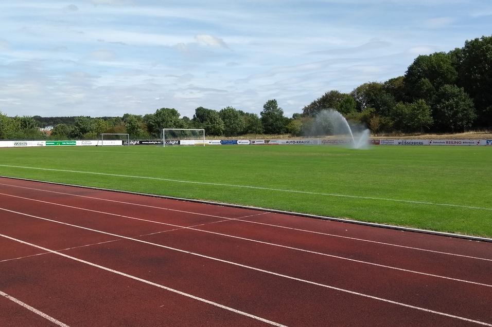 Gr&uuml;nes Fu&szlig;ballfeld mit einem Sprinkler, im Hintergrund eine Laufbahn und B&auml;ume unter einem blauen Himmel.