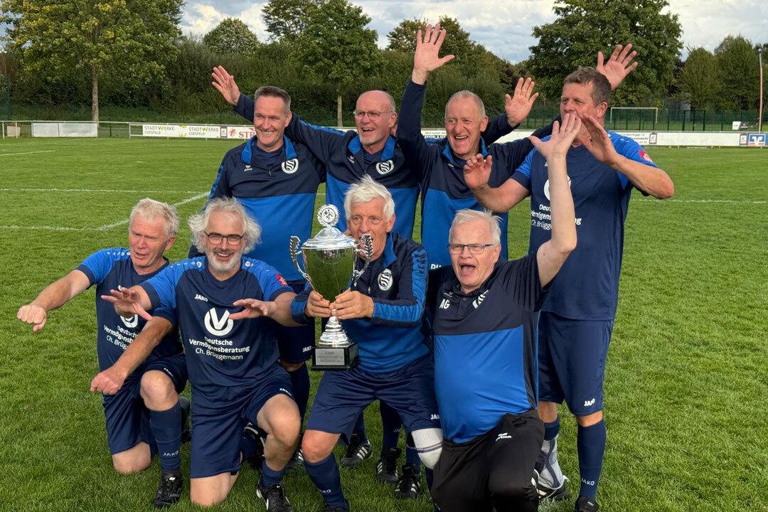 Gruppe von sieben Fu&szlig;ballspielern in blauen Trikots jubelt mit einem Pokal auf einem gr&uuml;nen Fu&szlig;ballfeld.