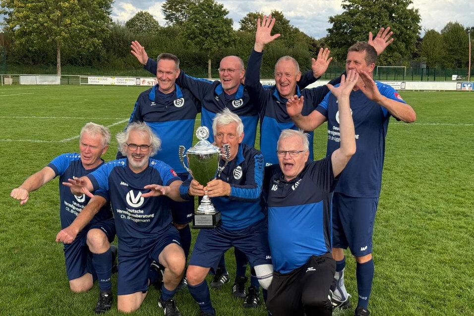 Gruppe von sieben Fußballspielern in blauen Trikots jubelt mit einem Pokal auf einem grünen Fußballfeld.
