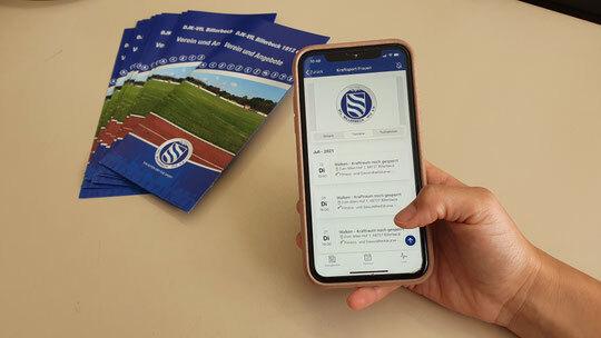 Eine Hand hält ein Smartphone mit einer App, während im Hintergrund Faltblätter mit Sportplatzmotiv liegen.