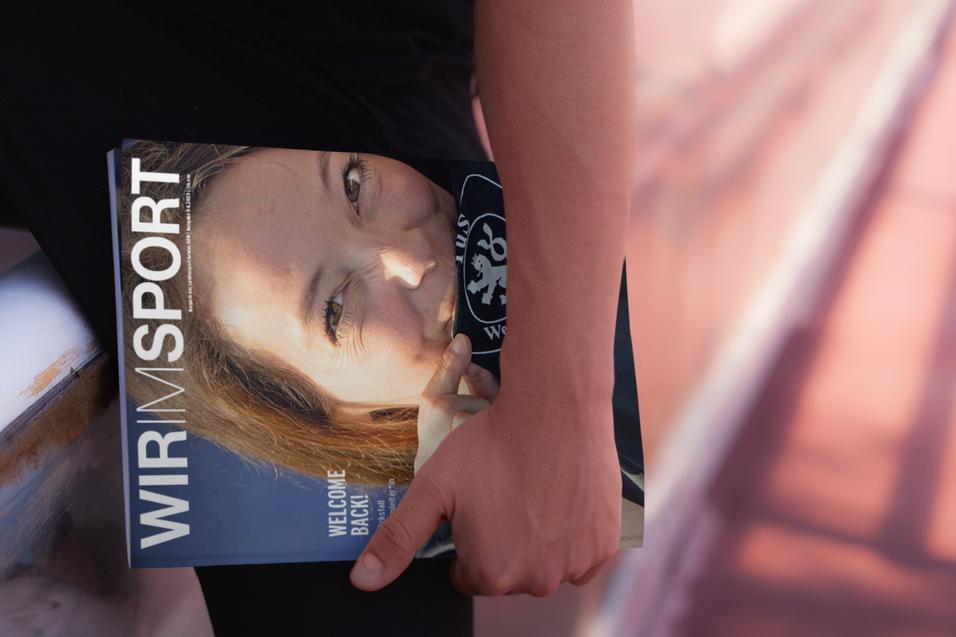 Nahaufnahme einer Person, die ein Magazin mit dem Titel "WIR IM SPORT" auf einem Sportgel&auml;nde h&auml;lt.