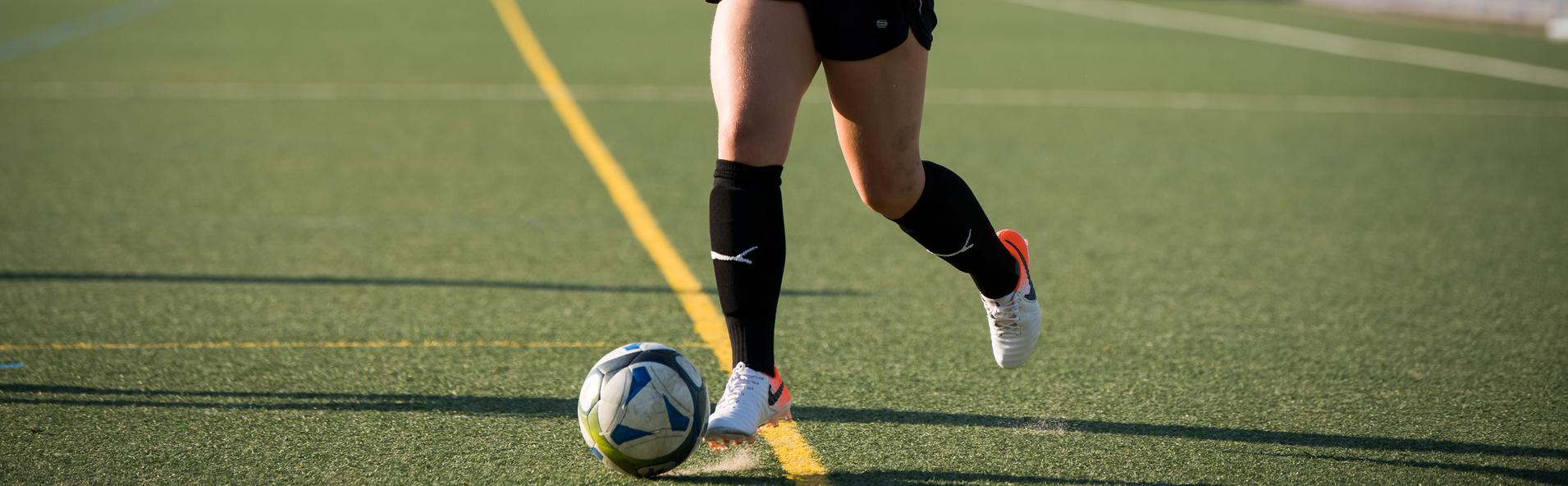 Eine Frau in schwarzer Sportkleidung dribbelt einen Fußball auf einem Kunstrasenplatz.