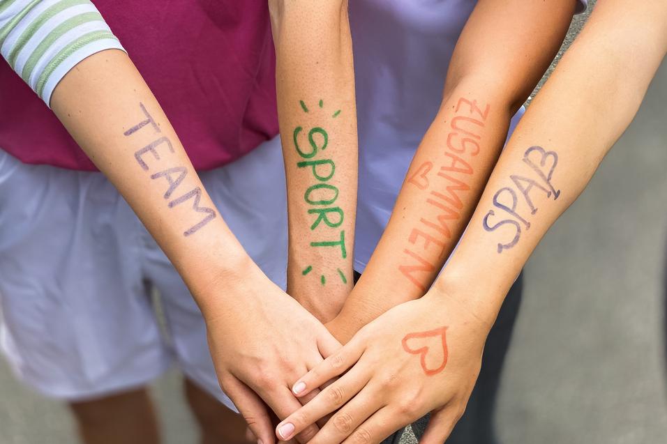 Vier Arme liegen übereinander, mit farbiger Schrift: „TEAM“, „SPORT“, „ZUSAMMEN“, „SPASS“ auf der Haut.