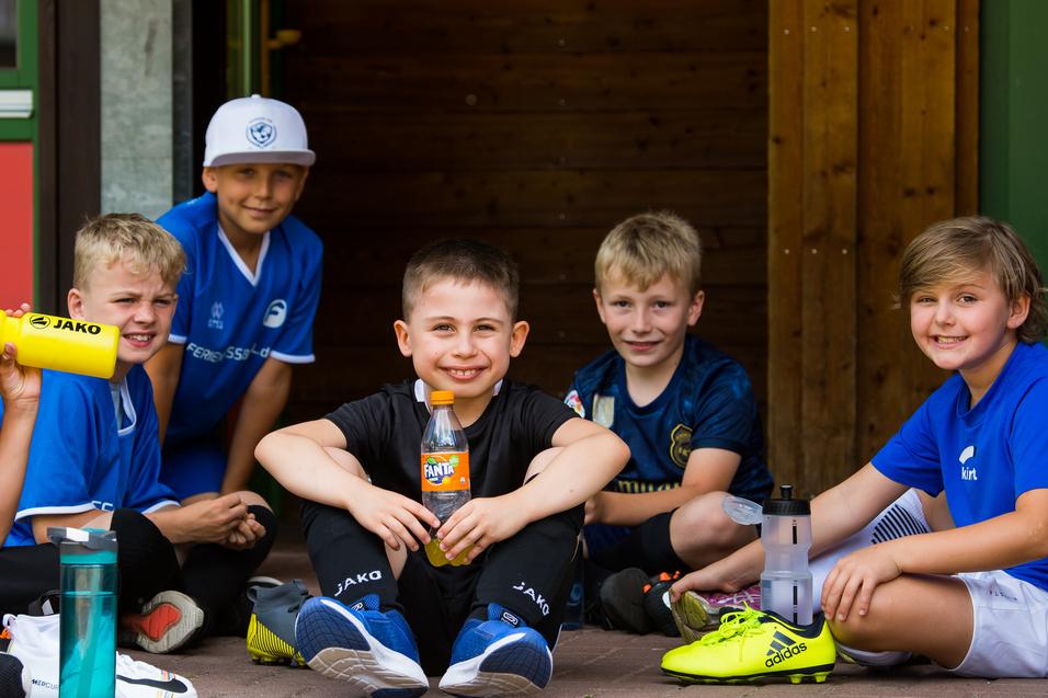 Sechs Kinder sitzen in Sportkleidung auf dem Boden, lächeln und halten Trinkflaschen und Snacks.
