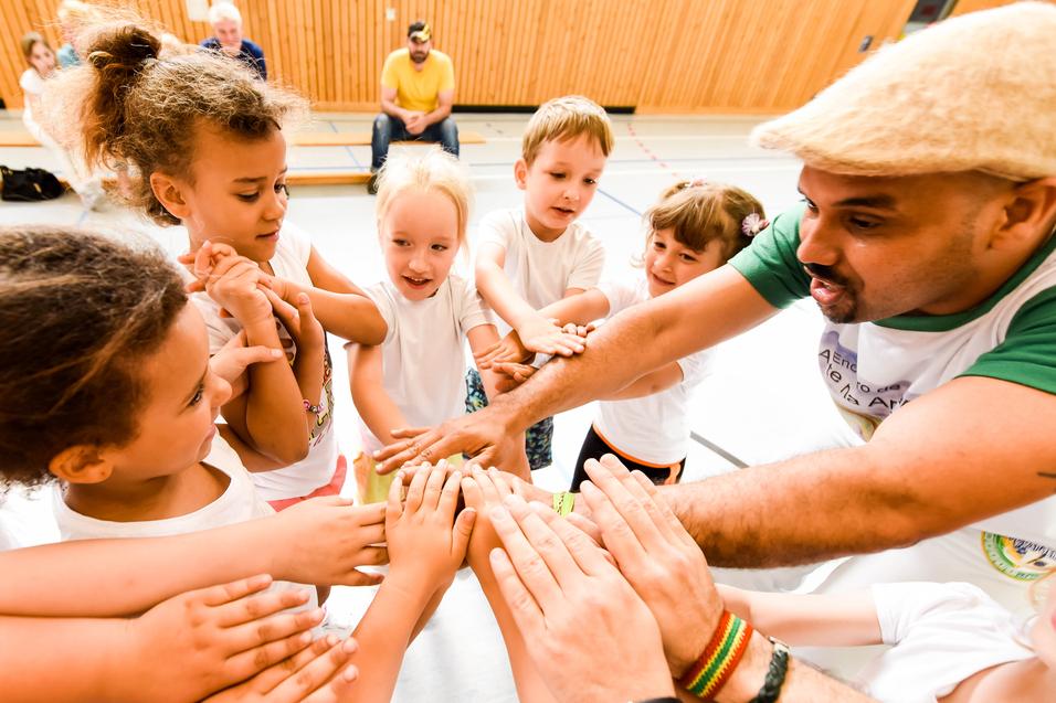 Gruppenspiel mit Kindern, die ihre Hände zum gemeinsamen Aufeinandertreffen heben, unter Anleitung eines Erwachsenen.
