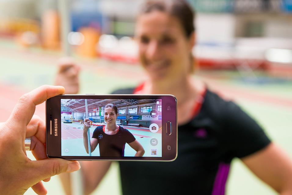 Hand hält ein Smartphone, das ein Porträt einer lächelnden Athletin mit Stab in einer Sporthalle zeigt.