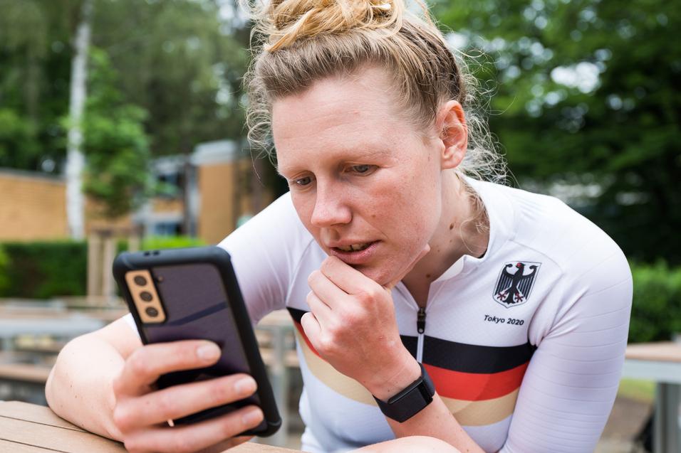 Athletin im weißen Trikot mit schwarzem und rotem Streifen schaut konzentriert auf ihr Smartphone in einer Außenanlage.