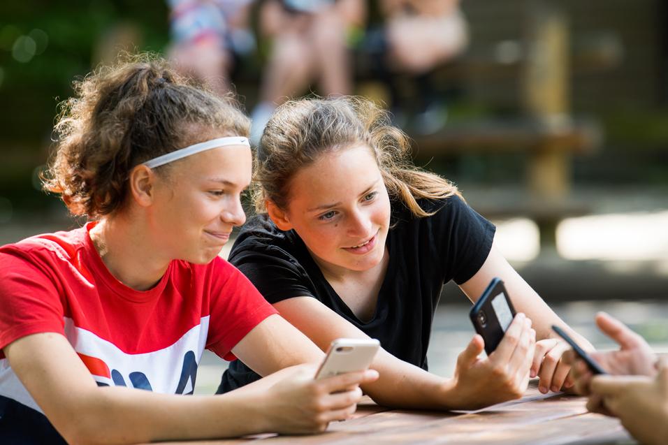 Zwei Mädchen sitzen an einem Tisch, schauen auf ihre Smartphones und lächeln dabei. Im Hintergrund sind weitere Personen sichtbar.