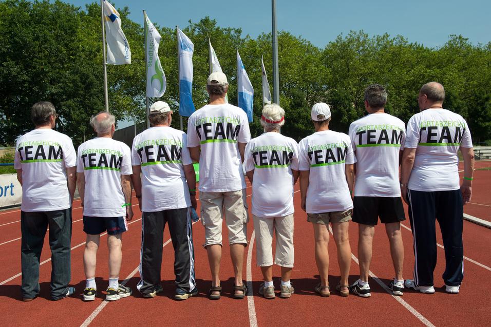 Gruppe von sieben Personen in weißen T-Shirts mit dem Aufdruck "TEAM" auf einer Laufbahn, Blick von hinten.